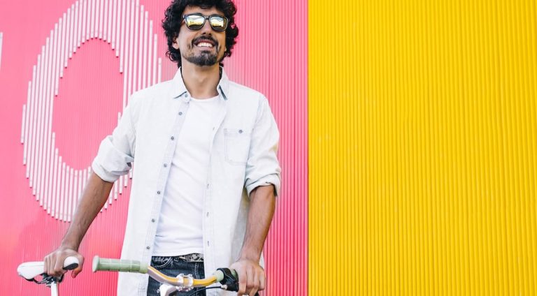 A man smiling, wearing sunglasses and holding on to a bicycling, standing in front of a brightly colored wall.