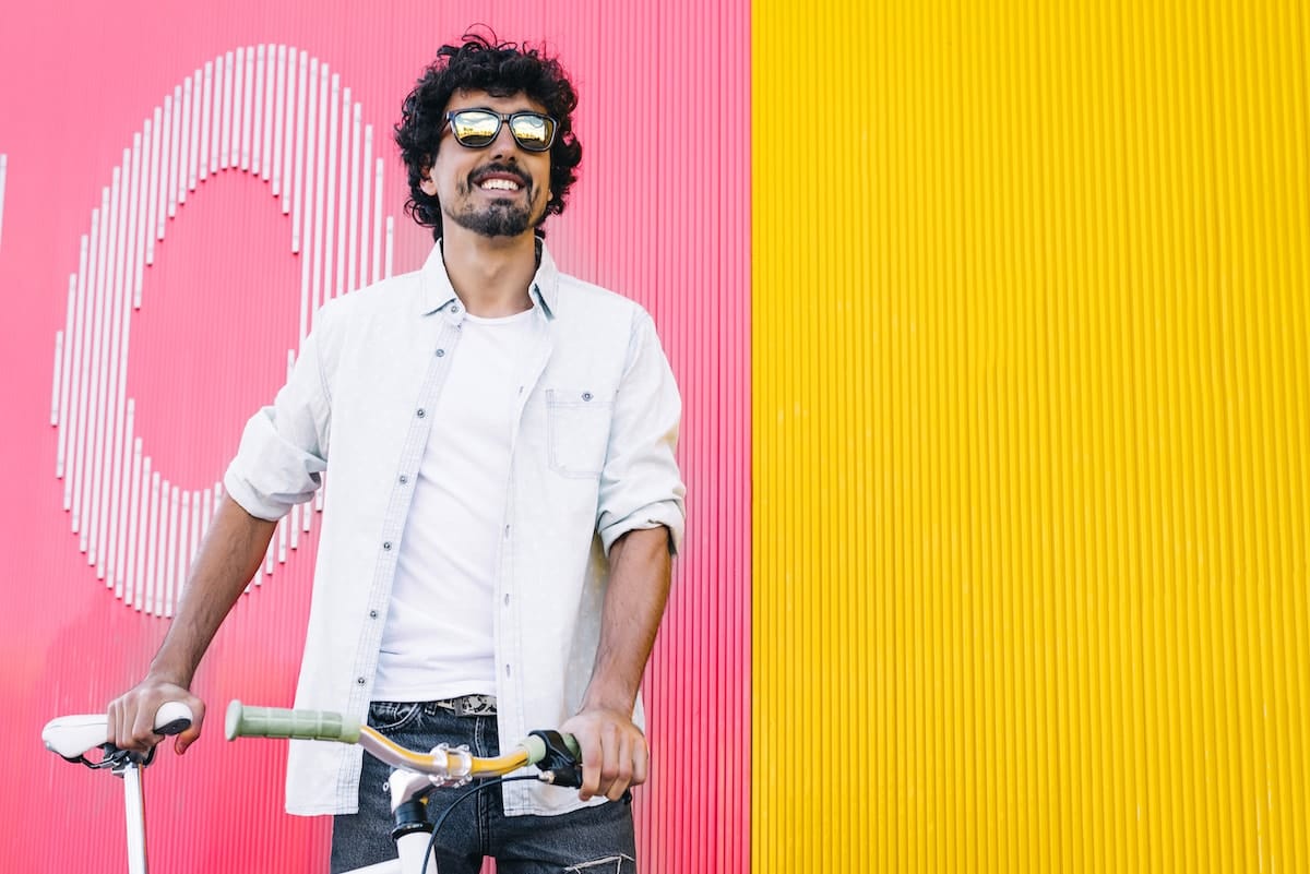 A man smiling, wearing sunglasses and holding on to a bicycling, standing in front of a brightly colored wall.