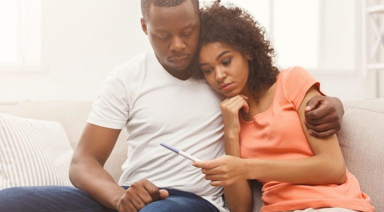 Black couple, seated next to one another on the couch, looking concerned at a pregnancy test.