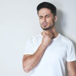 Young man having sore throat and touching his neck, wearing a loose white t-shirt against light grey background. Having difficulty swallowing.